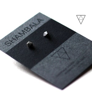 Puede incluir: Un par de pendientes de plata geométricos en una tarjeta negra con la palabra "SHAMBHALA" impresa en ella.