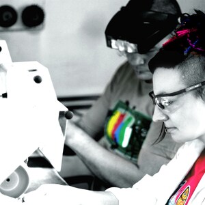 May include: A woman with pink and blue dreadlocks wears safety glasses and a white lab coat over a pink shirt. She is working with a white machine.