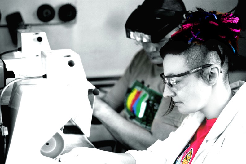 May include: A woman with pink and blue dreadlocks wears safety glasses and a white lab coat over a pink shirt. She is working with a white machine.
