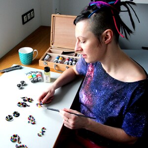 May include: A person paints colorful, rainbow-colored, polymer clay earrings with a small paintbrush. The earrings are shaped like swirls. The person is wearing a galaxy print shirt and has colorful dreadlocks.