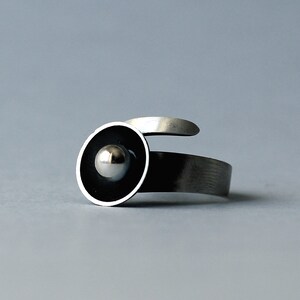 May include: A silver ring featuring a black circle with a silver sphere at its centre.