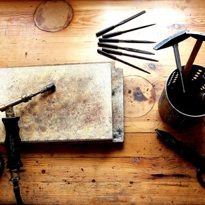 May include: A wooden workbench with a variety of tools for jewellery making, including a torch, files, hammers, and a small anvil.