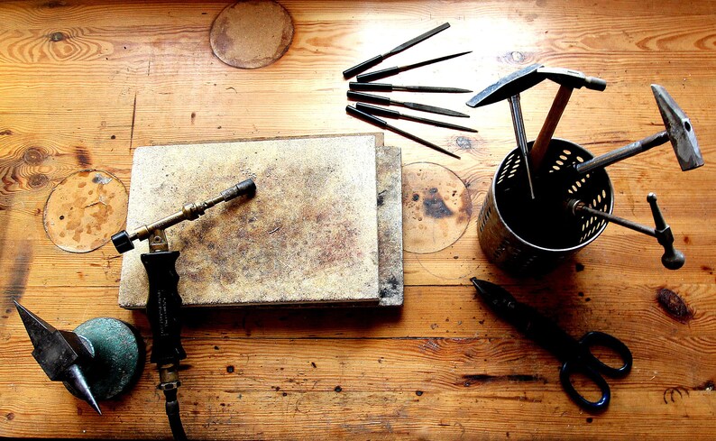 May include: A wooden workbench with a variety of tools for jewellery making, including a torch, files, hammers, and a small anvil.