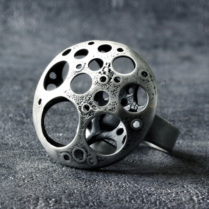 May include: A silver ring with a large, round, openwork design. The ring is made of metal and has a textured surface. The design features a series of circles and ovals, creating a unique and modern look.