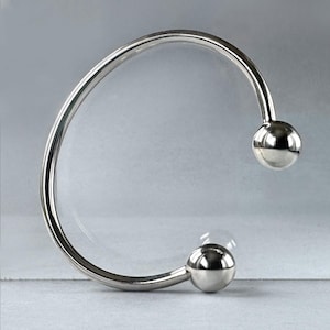 May include: A polished silver bracelet with a minimalist design. The open cuff features a smooth, curved band that terminates in two spherical accents. The bracelet's reflective surface catches the light, highlighting its sleek, modern aesthetic.