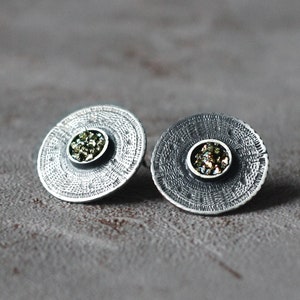 Op de afbeelding: Een paar zilveren oorbellen met een getextureerd rond ontwerp. Elke oorbel heeft een klein, rond, groen en gouden edelsteentje in het midden.