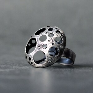 May include: A silver ring with a large, round, openwork design. The design features a pattern of circles and ovals, creating a textured and abstract look.