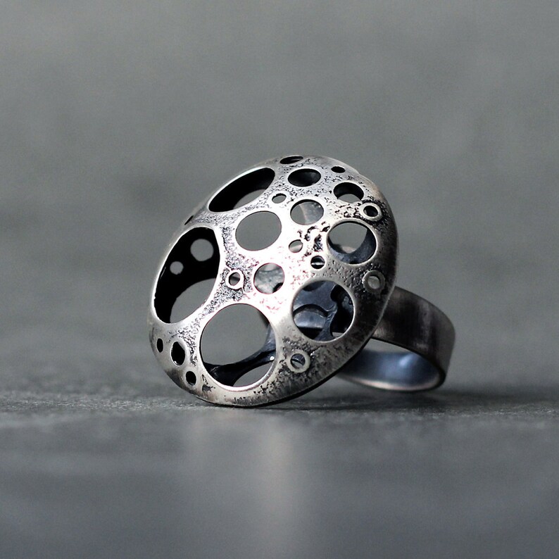 May include: A silver ring with a large, round, openwork design. The design features a pattern of circles and ovals, creating a textured and abstract look.