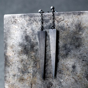 May include: A pair of silver earrings with long, thin, black chains and triangular pendants. The earrings are hanging from a grey, textured surface.