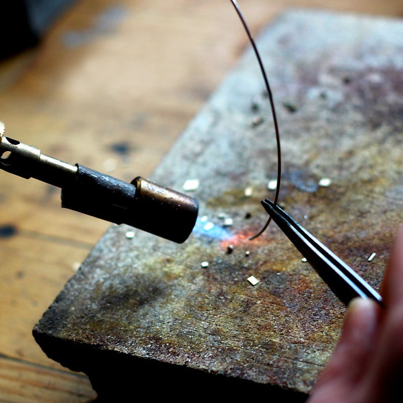 May include: A metal wire is being heated with a torch and held in place with tweezers on a metal surface.