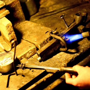 May include: A close-up of a metalworking torch with a blue flame being used to melt metal in a crucible. The torch is held by a hand, and the crucible is sitting on a wooden workbench.