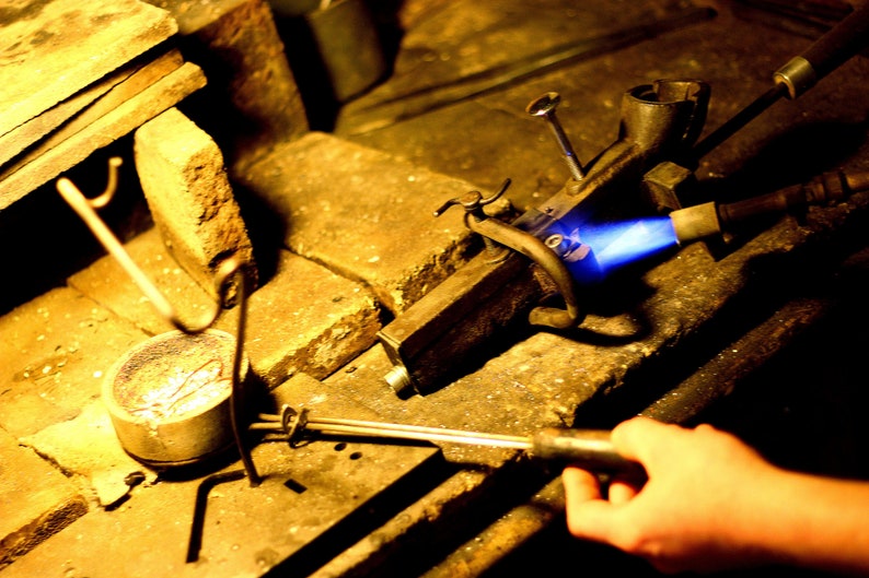 May include: A close-up of a metalworking torch with a blue flame being used to melt metal in a crucible. The torch is held by a hand, and the crucible is sitting on a wooden workbench.
