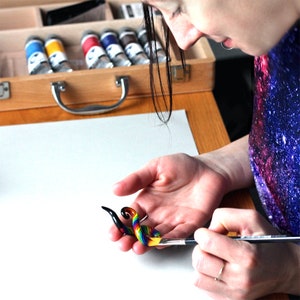 May include: A person paints a rainbow colored glass ear gauge with a fine paintbrush. The gauge is shaped like a spiral and has a black base.
