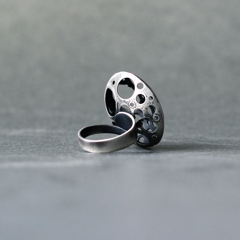 May include: A silver ring with a large, round, openwork design. The ring is made of metal and has a textured surface.