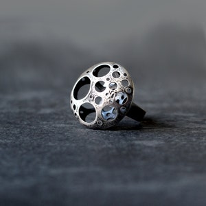 May include: A silver ring with a circular design featuring a pattern of small circles and holes. The ring is made of metal and has a textured surface.