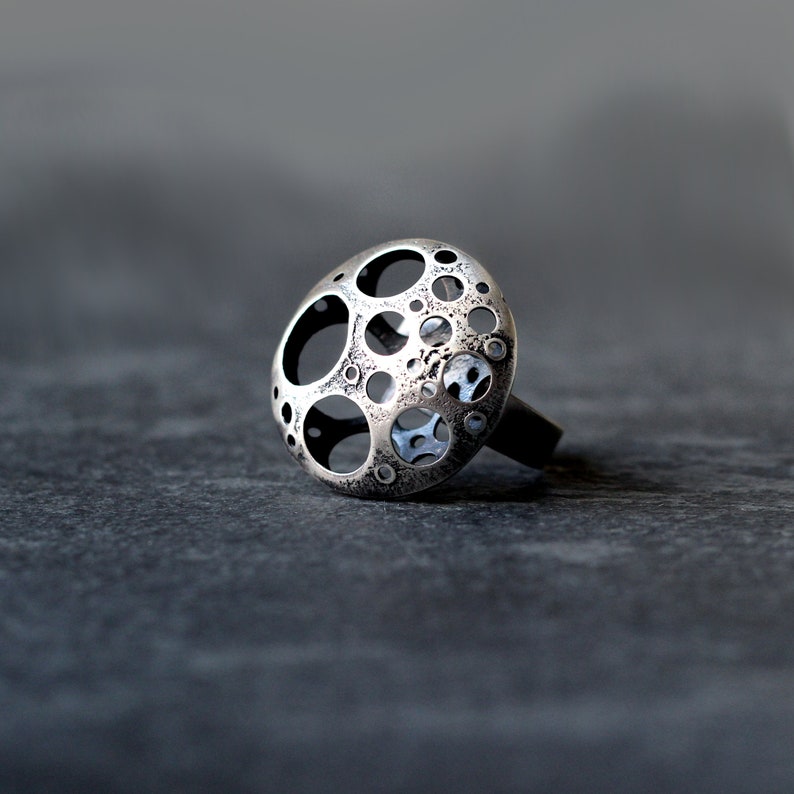 May include: A silver ring with a circular design featuring a pattern of small circles and holes. The ring is made of metal and has a textured surface.