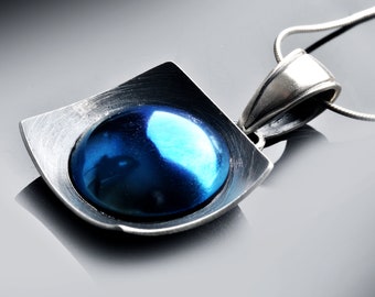 May include: A silver pendant with a square frame and a round blue glass cabochon. The pendant is attached to a silver chain.