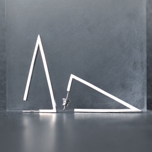 Könnte beinhalten: Ein Paar silberne Dreiecksohrringe. Die Ohrringe sind aus dünnem Metall gefertigt und haben ein schlichtes, geometrisches Design.