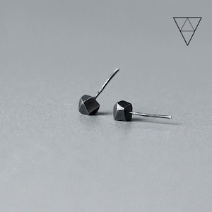 May include: A pair of small, dark gray stud earrings. Each earring features a faceted, geometric design. The posts are thin and silver-colored. The earrings are displayed on a light gray surface.