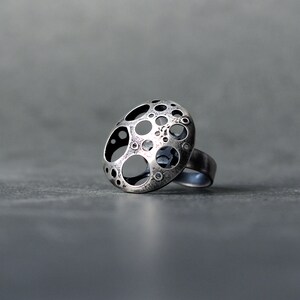 May include: A silver ring with a unique design featuring a circular pattern of cut-out circles. The ring is made of metal and has a textured finish.