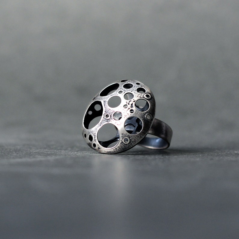 May include: A silver ring with a unique design featuring a circular pattern of cut-out circles. The ring is made of metal and has a textured finish.