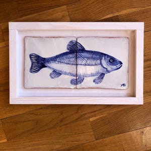 May include: A framed artwork featuring a blue and white fish illustration. The fish is depicted in a detailed, hand-drawn style, split across two rectangular tiles. The frame is made of light-colored wood, and the artwork is displayed on a wooden surface.