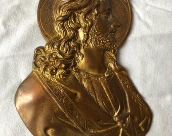 May include: A gold-toned metal wall plaque depicting a relief sculpture of Jesus Christ. The sculpture shows Jesus's head and shoulders, with a halo behind his head. The sculpture is detailed and intricate, with a textured surface.