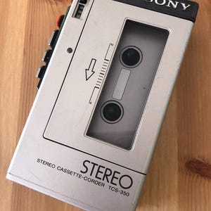 May include: A vintage silver Sony Stereo Cassette-Corder TCS-350. The portable audio device features a cassette tape window, control buttons, and the brand name "SONY" in black. The device is on a wooden surface.