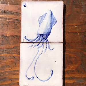 May include: Two square ceramic tiles with a hand-painted blue squid design on a white background. The squid is depicted with detailed tentacles and a stylized form. A small blue symbol is in the upper left corner of one tile. The tiles are set against a wooden surface.