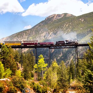 May include: A red and yellow train travels across a metal bridge over a rocky riverbed. The train is surrounded by lush green trees and mountains in the background. The sky is blue with white clouds.