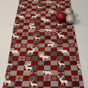 Peut inclure: Un chemin de table à carreaux rouges et gris avec un motif de rennes et d'élans blancs. Le chemin de table est décoré d'ornements de Noël rouges et argentés.