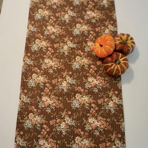 May include: A brown floral table runner with vertical stripes. The runner is decorated with a pattern of pink and blue flowers. Three small orange pumpkins are placed on the runner.