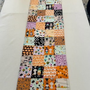 May include: A rectangular Halloween table runner. The runner is made of patchwork squares in various colors, including orange, black, purple, and white. The squares feature Halloween-related designs such as pumpkins, stars, and ghosts. The runner is approximately 152 cm long.