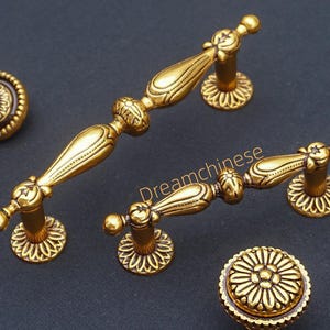 Peut inclure: Ensemble de quincaillerie de meubles en laiton antique, comprenant des poignées et un bouton. Le matériel présente des détails floraux ornés et une finition dorée chaude. Le texte "Dreamchinese" est visible.