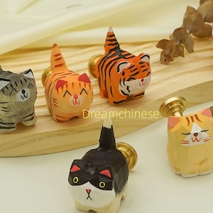 Könnte beinhalten: Eine Sammlung handgeschnitzter Schrankknöpfe in Katzenform aus Holz. Die Knöpfe weisen verschiedene Katzendesigns auf, darunter Tabby-, Tigerstreifen- und Schildpattmuster, mit Messingbeschlägen. Die Knöpfe werden auf einer Holzoberfläche präsentiert.