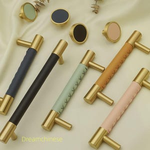 Pode incluir: Uma coleção de puxadores e puxadores de armário em latão com detalhes em couro ou semelhantes em várias cores, incluindo azul, preto, verde menta, castanho e rosa. Os puxadores e puxadores estão dispostos sobre um tecido creme.