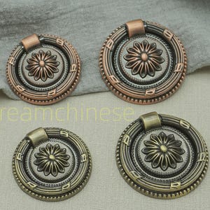 Peut inclure: Ensemble de six poignées de tiroir de style antique de différentes tailles. Elles présentent un motif floral central et sont finies en bronze, cuivre et laiton. Le texte "Dreamchinese" est visible.