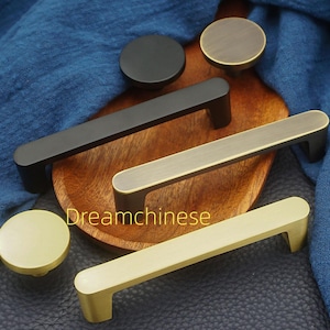 May include: A selection of modern cabinet hardware in various finishes. The image showcases black, brass, and gold-toned handles and knobs. The hardware is displayed on a wooden tray and a dark blue surface.