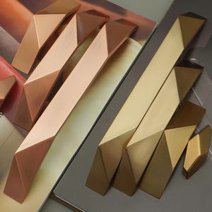 Könnte beinhalten: Geometrische Schrankbeschläge in Kupfer- und Goldtönen. Das Bild zeigt mehrere Schrankgriffe und -knöpfe mit einem facettierten, eckigen Design. Die Beschläge sind auf einem neutralen Hintergrund angeordnet, wodurch die metallischen Oberflächen und die moderne Ästhetik hervorgehoben werden.
