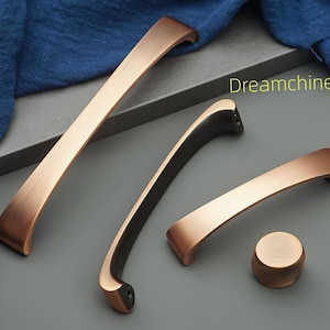 May include: A selection of copper-toned cabinet handles and a knob. The set includes two curved handles, one straight handle, and a round knob. The items are arranged on a grey surface, with a blue fabric in the background. The text "Dreamchinese" is visible.