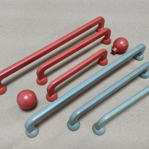 以下が含まれることがあります： 赤と青緑色の取っ手とノブを含む、キャビネットハードウェアのコレクション。取っ手は長方形で端が丸く、ノブは球形です。さまざまなキャビネットの寸法に対応する、さまざまなサイズがあります。