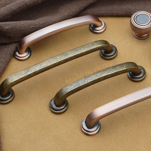 以下が含まれることがあります： ブロンズとアンティーク真鍮のキャビネットハードウェアのコレクション。カーブしたハンドルと丸いノブが含まれています。ハードウェアはヴィンテージの美学を持ち、さまざまな仕上げとスタイルがあります。画像には「Made by Henry」というテキストが含まれています。