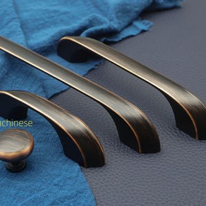 May include: Bronze-finished cabinet hardware set, including three pull handles and one knob. The handles have a curved design and are set against a blue fabric and a dark blue surface.