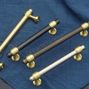 May include: Four cabinet pulls in gold, black, bronze, and silver. Each pull has a cylindrical bar with gold accents at the ends and a matching gold base. The pulls are arranged on a blue fabric background.