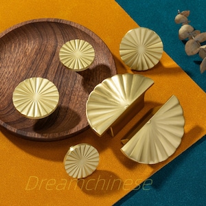 May include: A collection of gold-colored cabinet knobs and pulls with a fan-like design. The knobs are round, while the pulls are semi-circular. They are arranged on a wooden tray and a colorful background. The text "Dreamchinese" is visible.