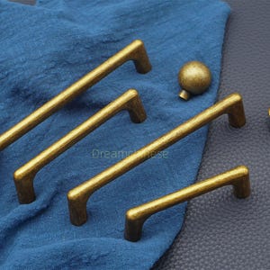 May include: A collection of antique gold-colored cabinet hardware, including four pull handles of varying sizes and a round knob. The hardware is displayed on a blue cloth and a dark gray surface, showcasing a textured finish.