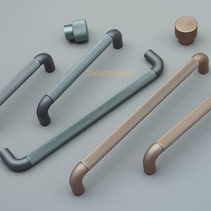 May include: A collection of cabinet hardware in various colors, including gray, blue-gray, and bronze. The handles and knobs have a textured, diamond-patterned grip. The hardware is displayed on a light gray surface.
