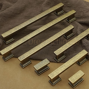 Peut inclure: Ensemble de poignées et de boutons de meubles en laiton vieilli. Les poignées sont rectangulaires avec une finition légèrement vieillie, et les boutons sont carrés. Le mot "Dreamchine" est imprimé. Parfait pour la restauration de meubles.