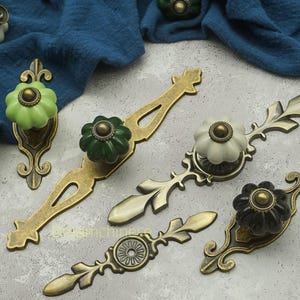Op de afbeelding: Een verzameling decoratieve kastbeslagen met bloemvormige keramische knoppen in groen, wit en zwart. De knoppen zijn gemonteerd op sierlijke, antiek messingkleurige achterplaten en handgrepen, met een vintage uitstraling.