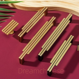 May include: A selection of gold-coloured cabinet handles and a knob, displayed on a burgundy surface. The handles have a linear design with vertical grooves. The text "Dreamcheese" is visible at the bottom. The hardware is set against a wooden backdrop.